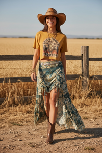 Woman in a yellow rodeo shirt and patterned skirt standing in a field with a wooden fence wearing cowboy boots.